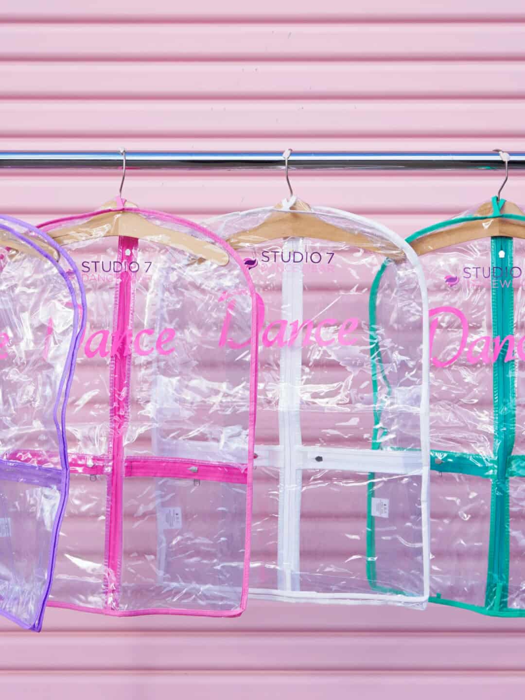 Clear garment bags with pink and green zippers on hangers against a pink background.