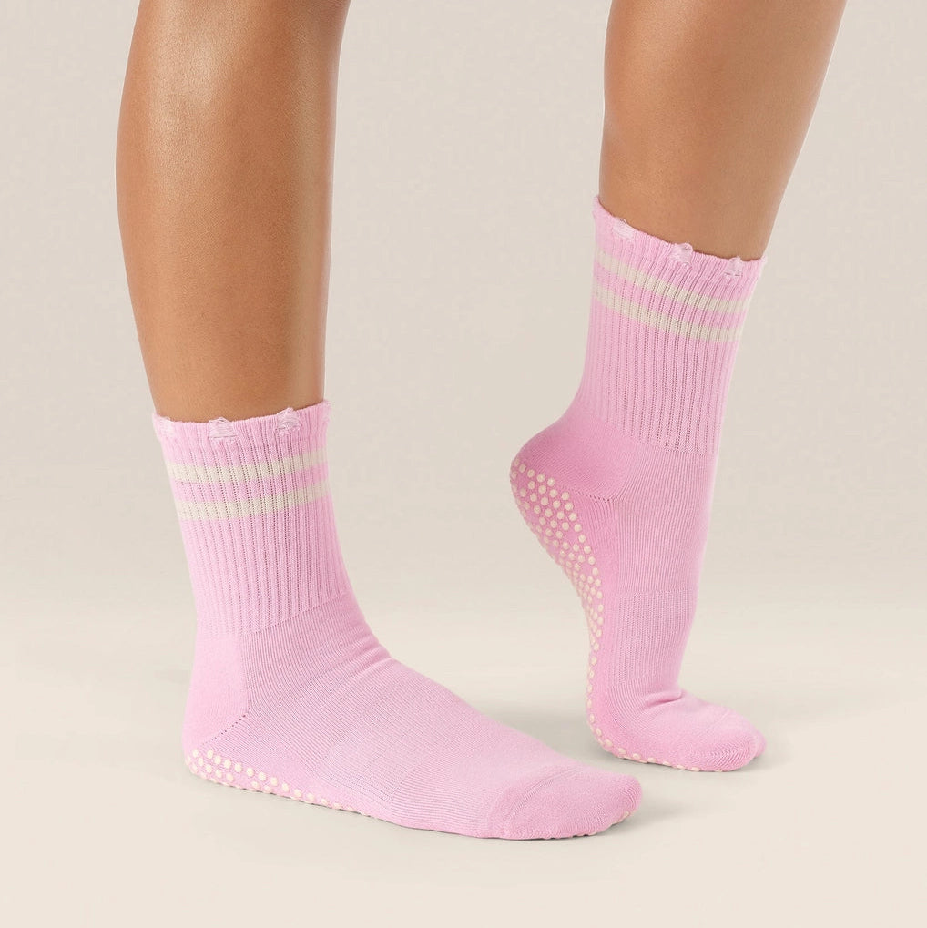 A pair of light pink crew socks with grip dots on the sole and a subtle pale green stripe detail near the cuff, shown on a model's feet against a neutral beige background.