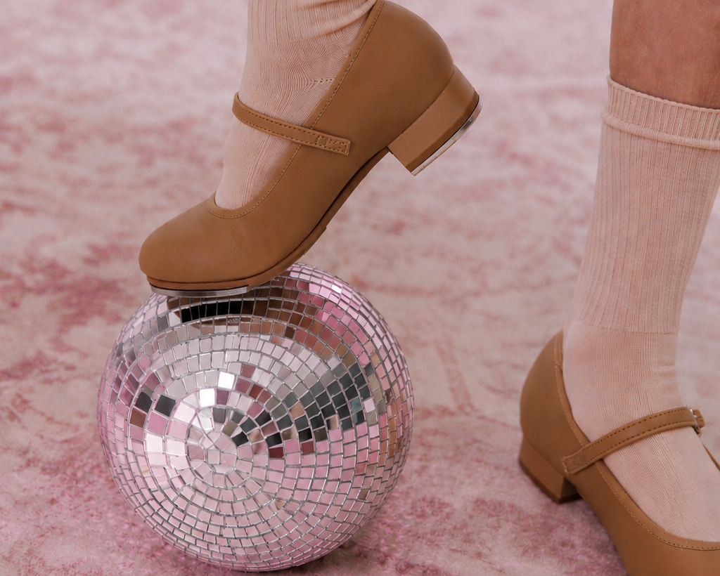 A person wearing beige Mary Jane low-heel tap shoes and ribbed beige socks stands on a pink patterned rug, with one foot resting on a mirrored disco ball.