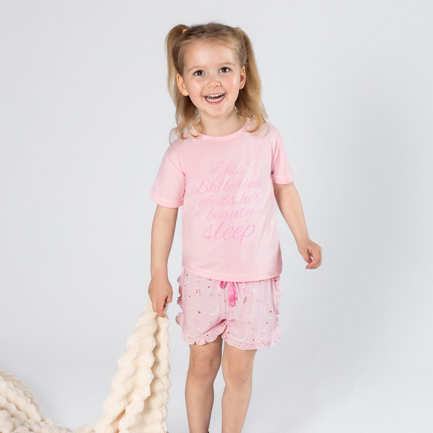 Young girl in pink outfit standing next to a cream blanket on a white background