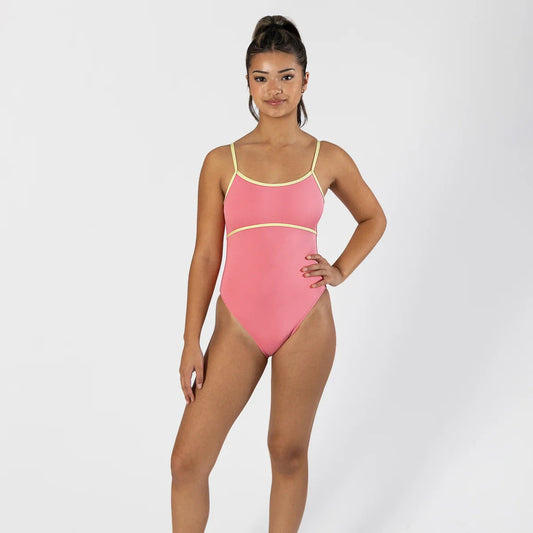 A woman modeling a pink one-piece swimsuit with thin yellow straps and trim, paired with white socks and sneakers, against a light gray studio background.
