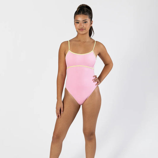 A woman modeling a light pink one-piece swimsuit with thin yellow straps and a thin yellow waistband, paired with white socks and white sneakers, against a light gray studio background.