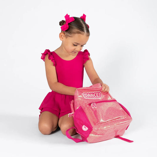 A young girl in a bright pink leotard and matching hair bows kneels on a white studio floor, looking into a pink glittery dance backpack with the brand name 'DANCE!' visible on the front.