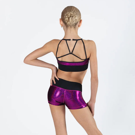 A young gymnast is shown from the back wearing a black and purple sports bra and matching shiny purple shorts against a white studio background.